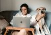 Kvinna som jobbar i soffan med en laptop ovanpå ett litet anpassat bord, gosar samtidigt med en hund.